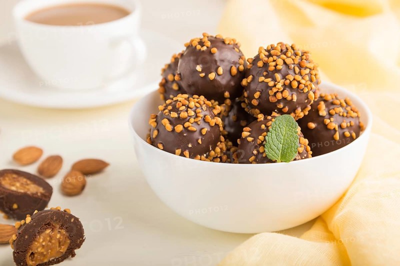 Chocolate caramel ball candies with almonds and a cup of coffee on a white wooden background and yellow textile. Side view, close up, selective focus
