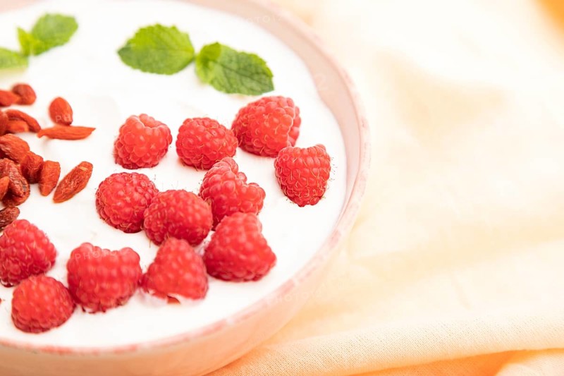 Yogurt with raspberry and goji berries in ceramic bowl on white concrete background and orange linen textile. Side view, close up, selective focus