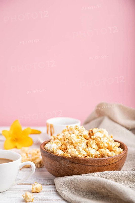 Popcorn with caramel in wooden bowl and a cup of coffee on a white and pink background and linen textile. Side view, copy space, selective focus