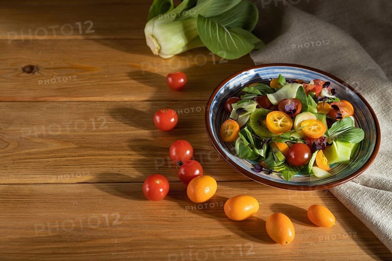 Vegetarian salad of pac choi cabbage, kiwi, tomatoes, kumquat, microgreen sprouts on a wooden background and linen textile. Hard light, contrast. Side view, selective focus, copy space