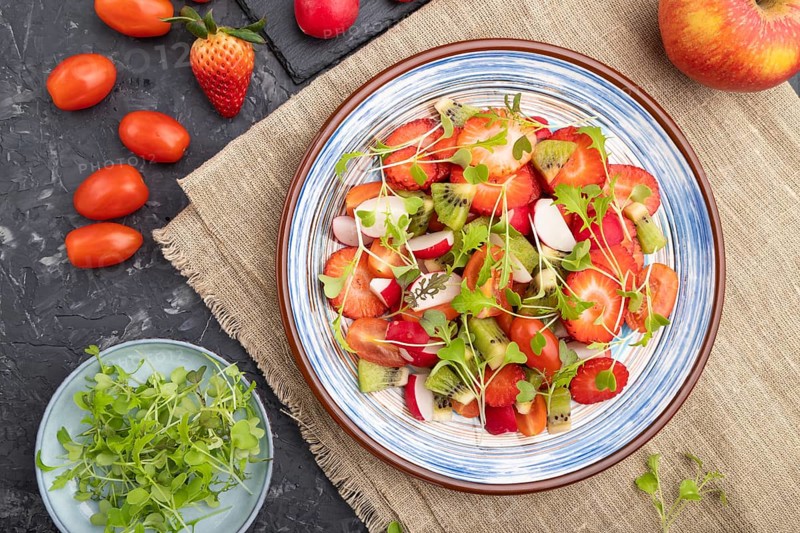 Vegetarian fruits and vegetables salad of strawberry, kiwi, tomatoes, microgreen sprouts on black concrete background and linen textile. Top view, flat lay, close up