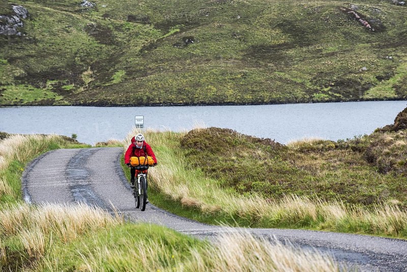 Lonely female biker cycling through the Scottish Highlands on heavily laden touring bicycle along single track road in bad rainy weather, Scotland, UK