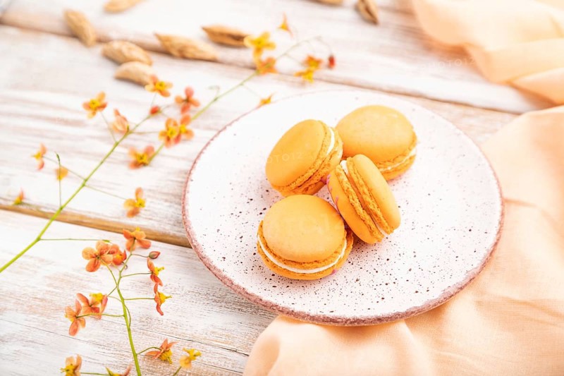Orange macarons or macaroons cakes with cup of apricot juice on a white wooden background and orange linen textile. Side view, close up, selective focus