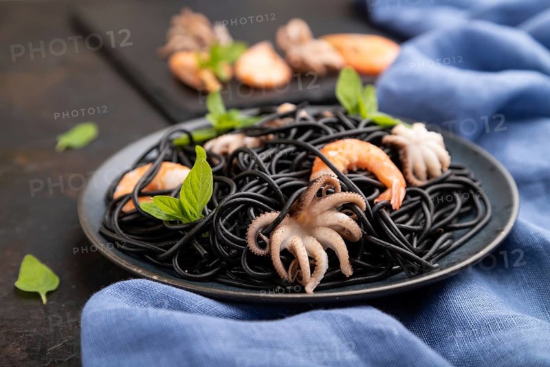 Black cuttlefish ink pasta with shrimps or prawns and small octopuses on black concrete background and blue textile. Side view, close up, selective focus