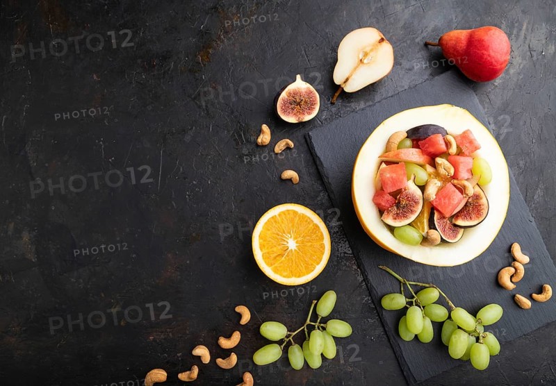 Vegetarian fruit salad of watermelon, grapes, figs, pear, orange, cashew on slate board on a black concrete background. Top view, flat lay, copy space