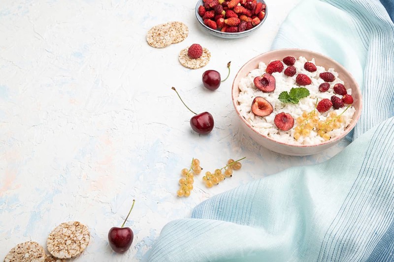 Rice flakes porridge with milk and strawberry in ceramic bowl on white concrete background and blue linen textile. Side view, copy space