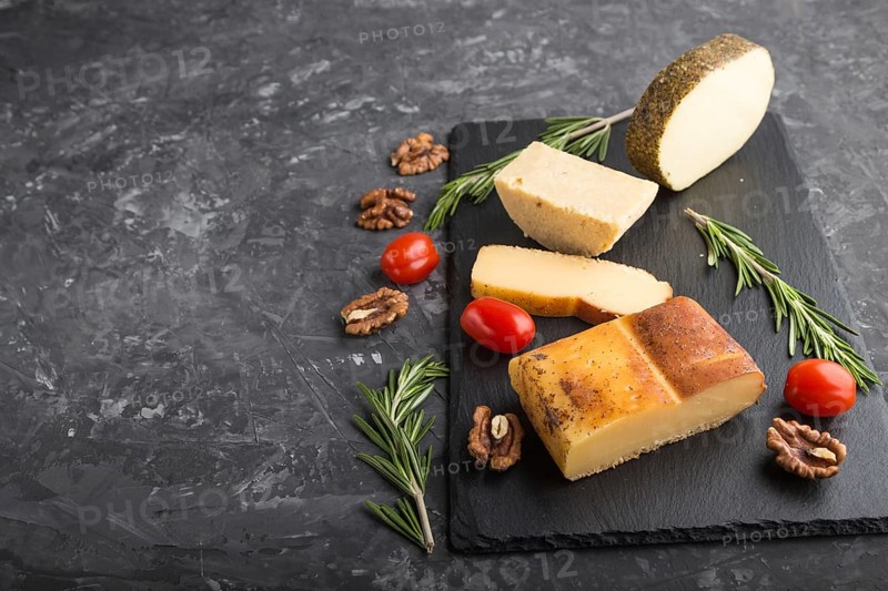 Smoked cheese and various types of cheese with rosemary and tomatoes on black slate board on a black concrete background. Side view, copy space, selective focus