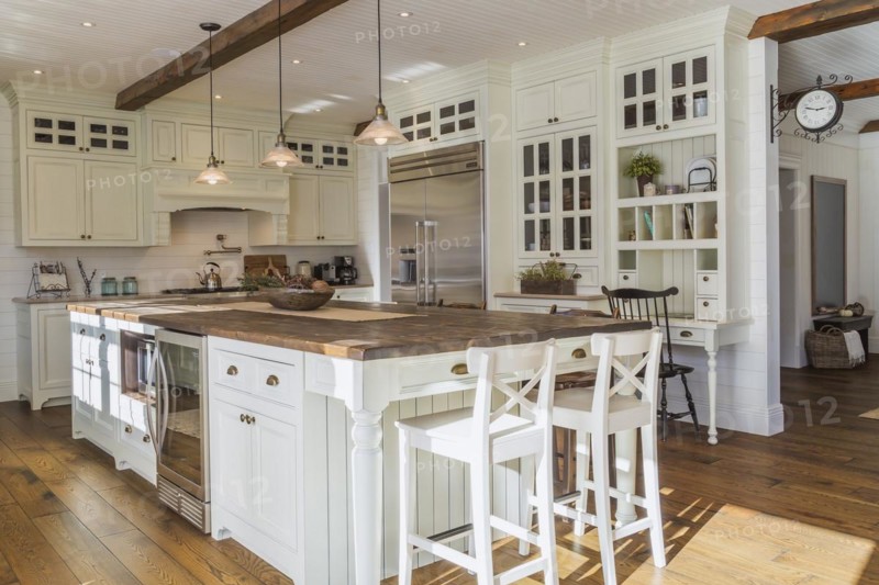 White painted maple wood antique style cabinets with tan coloured quartz countertops, island with brown stained wood plank countertop and high back bar stools in kitchen with stained oak wood floorboards inside country style home, Quebec, Canada, North America
