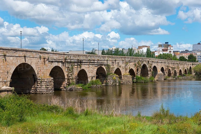 Long historic stone bridge over a river, surrounded by nature and buildings under a blue sky, Roman bridge, Puente Romano, arched bridge with wedge stone vault, Guadiana River, Mérida, Merida, Badajoz, Extremadura, Spain, Europe