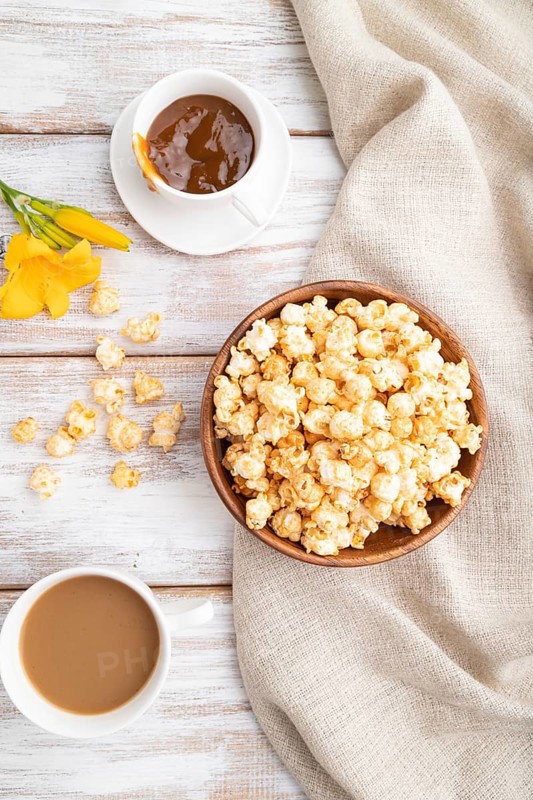 Popcorn with caramel in wooden bowl and a cup of coffee on a white wooden background and linen textile. Top view, flat lay, close up