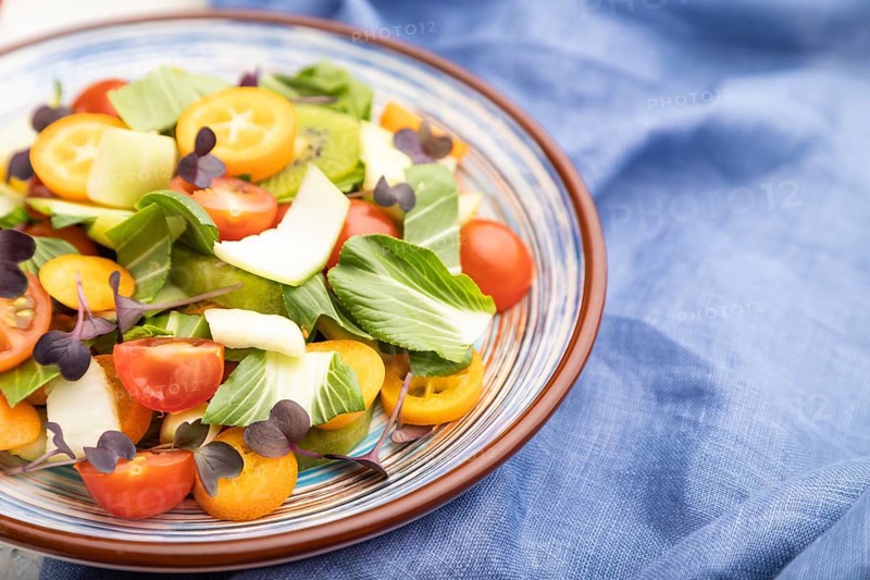 Vegetarian salad of pac choi cabbage, kiwi, tomatoes, kumquat, microgreen sprouts on a white concrete background and blue linen textile. Side view, close up, selective focus