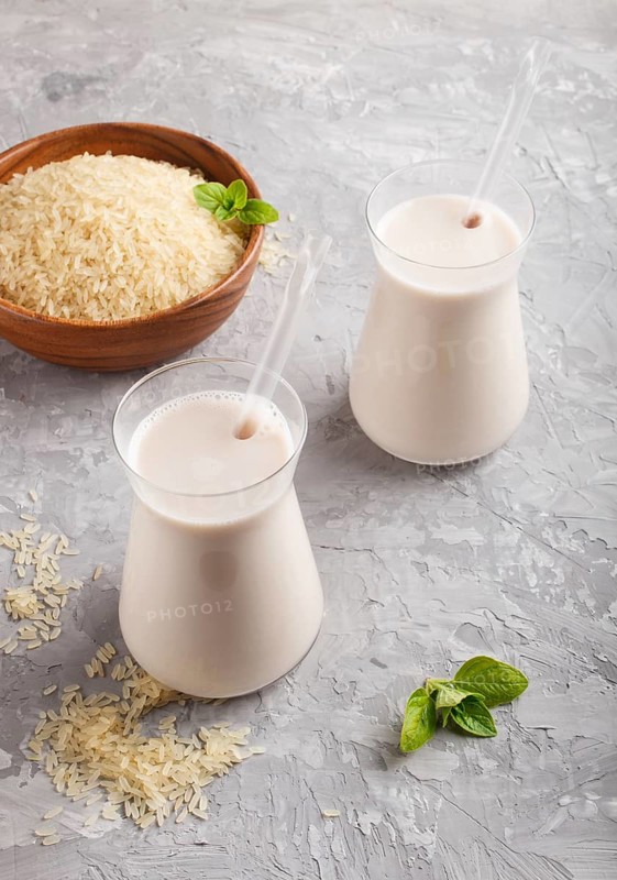 Organic non dairy rice milk in glass and wooden plate with rice seeds on a gray concrete background. Vegan healthy food concept, close up, side view