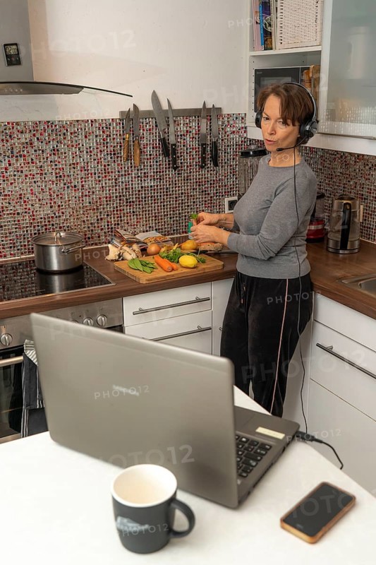 Woman, mid-50s, works from home, with laptop and communicates with colleagues via headset, home office, in the kitchen, while cooking