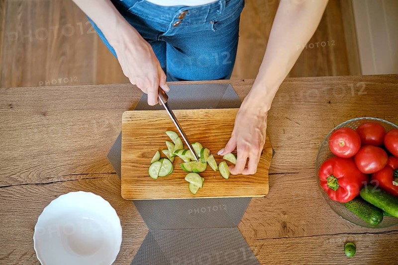 Overhead view of female hands slicing fresh cucumber on wooden chopping board