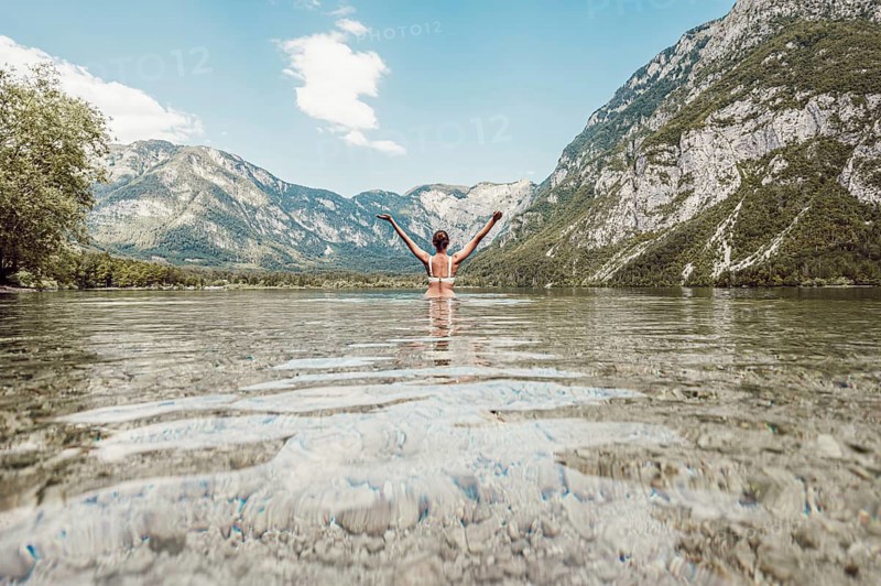 Woman with arms outstretched to the sky on both sides in a clear lake, surrounded by mountains and under a wide sky, Soca Valley, Slovenia, Europe