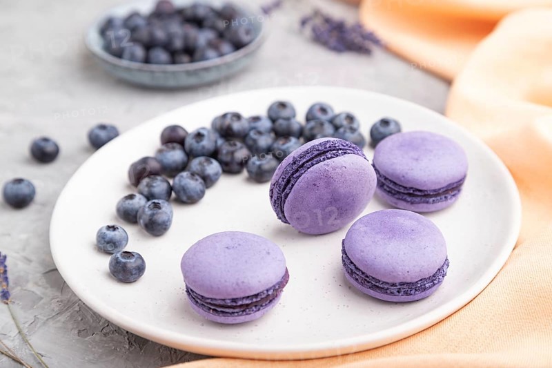 Purple macarons or macaroons cakes with blueberries on white ceramic plate on a gray concrete background and orange textile. Side view, close up, selective focus