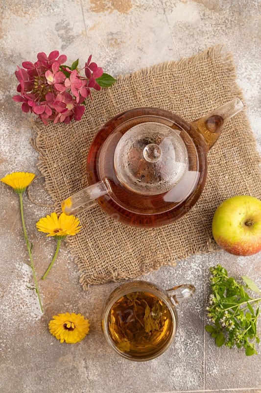 Red tea with herbs in glass teapot on brown concrete background and linen textile. Healthy drink concept. Top view, flat lay, close up