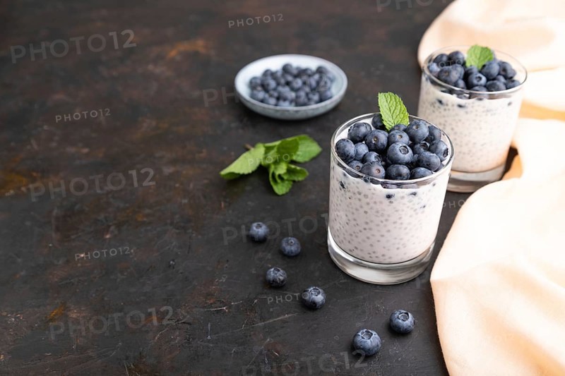 Yogurt with blueberry and chia in glass on black concrete background and orange linen textile. Side view, copy space