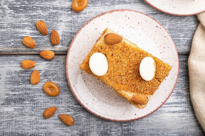 Honey cake with milk cream, caramel, almonds and a cup of coffee on a gray wooden background and linen textile. Top view, flat lay, close up