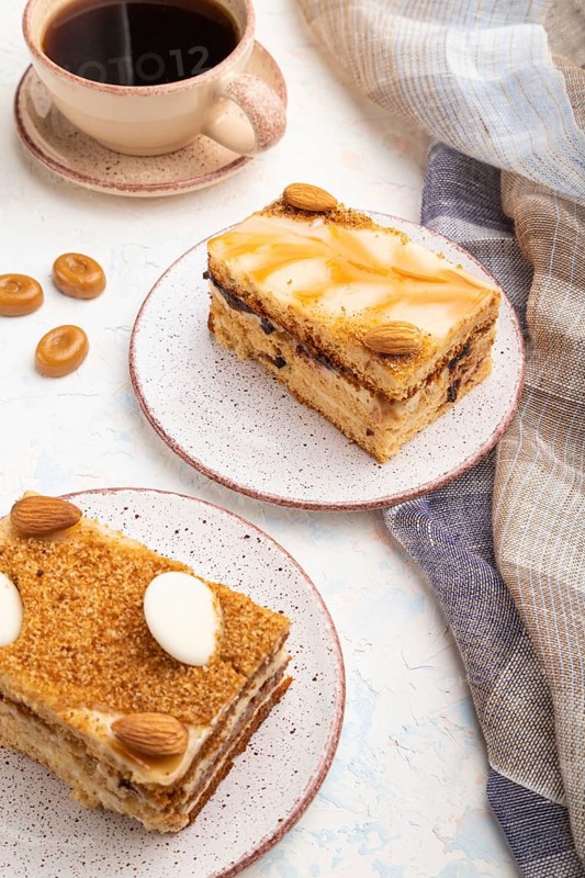 Honey cake with milk cream, caramel, almonds and a cup of coffee on a white concrete background and linen textile. Side view, close up