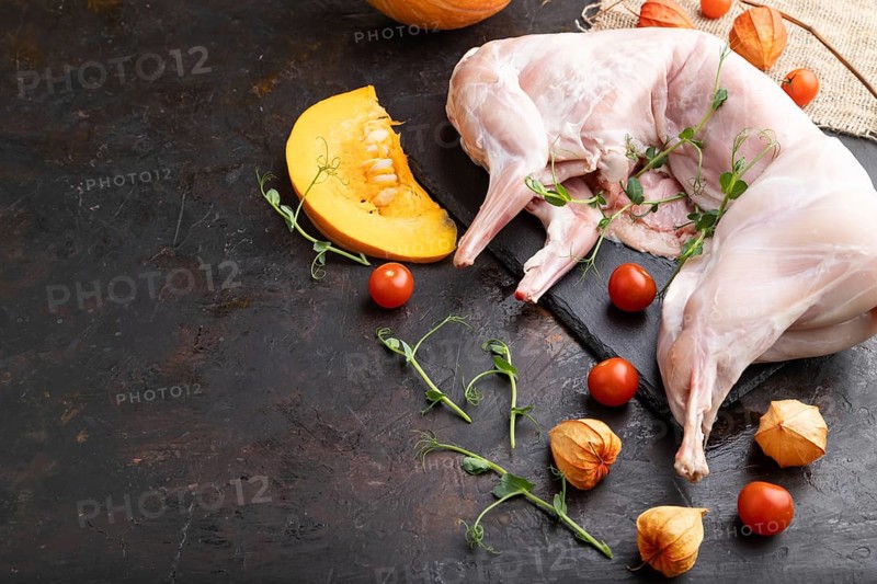 Whole raw rabbit with pumpkin, tomatoes, pea sprouts on a black concrete background and linen textile. Side view, copy space