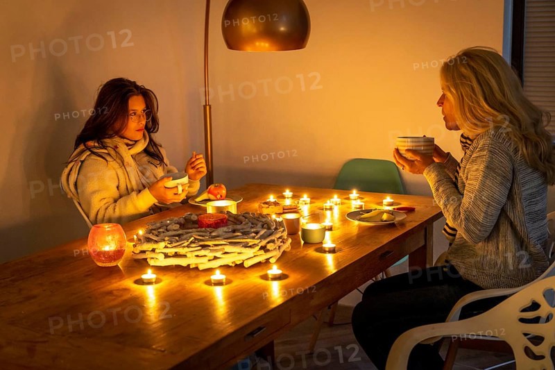 Symbolic image energy saving, cold winter, energy crisis, cold flat, mother and daughter dressed warmly at dinner, only by candlelight, heating on to a minimum