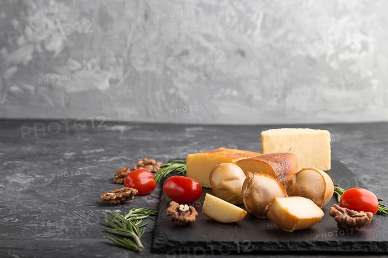 Smoked cheese and various types of cheese with rosemary and tomatoes on black slate board on a black concrete background. Side view, copy space, selective focus
