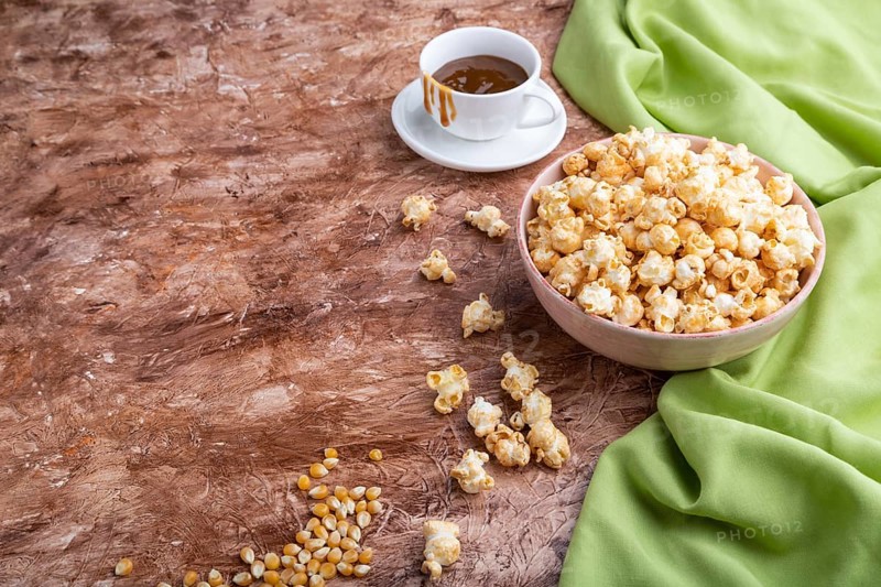Popcorn with caramel in ceramic bowl on brown concrete background and green textile. Side view, copy space