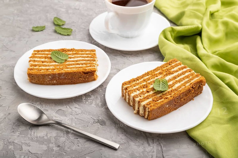 Homemade honey cake with milk cream and mint with cup of coffee on a gray concrete background and green textile. Side view, close up
