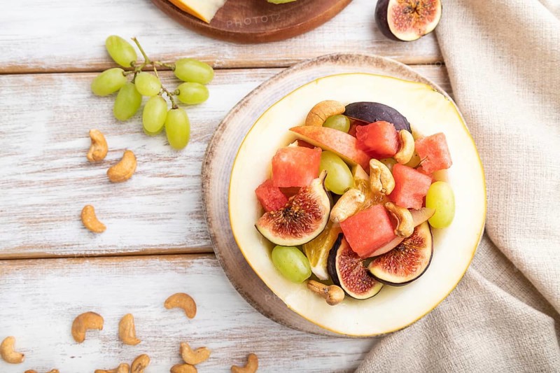 Vegetarian fruit salad of watermelon, grapes, figs, pear, orange, cashew on white wooden background and linen textile. Top view, flat lay, close up