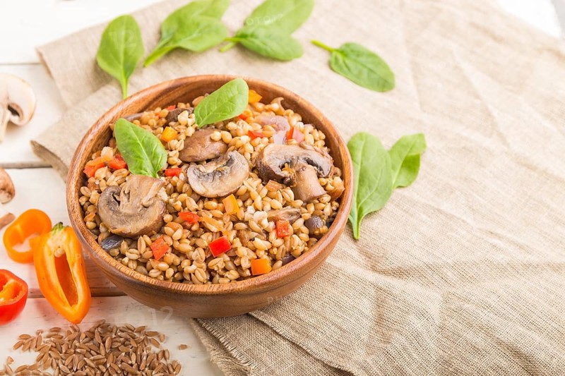 Spelt (dinkel wheat) porridge with vegetables and mushrooms in wooden bowl on a white wooden background and linen textile. Side view, selective focus. Russian traditional cuisine