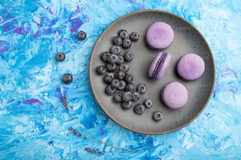 Purple macarons or macaroons cakes with blueberries on ceramic plate on a blue concrete background. Flat lay, top view, close up