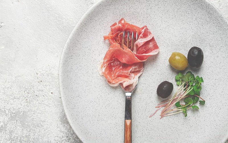 Prosciutto on a fork, top view, olives and micro-greens on a plate, light background, no people