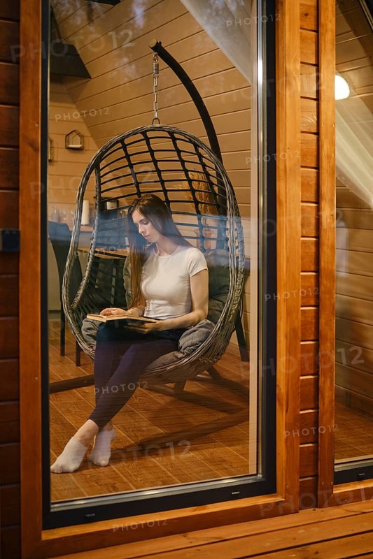 Young woman sitting in a cozy hanging chair, engrossed in reading a book. A woman spends her evening reading a novel