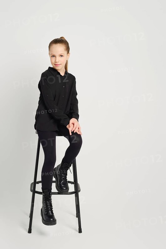 Young girl dressed in a black shirt and tights sits casually on a high stool against a white background