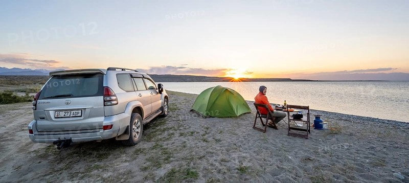 Camping at the lakeside, young man sitting next to tent on camping chair, having dinner, off-road vehicle, sunset at Issyk Kul Lake, Issyk Kul, Kyrgyzstan, Asia