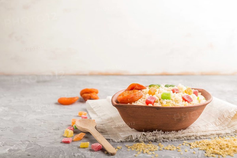 Bulgur porridge with dried apricots and candied fruits in clay bowl on a gray concrete background and linen textile. Side view, copy space, selective focus. Turkish traditional cuisine