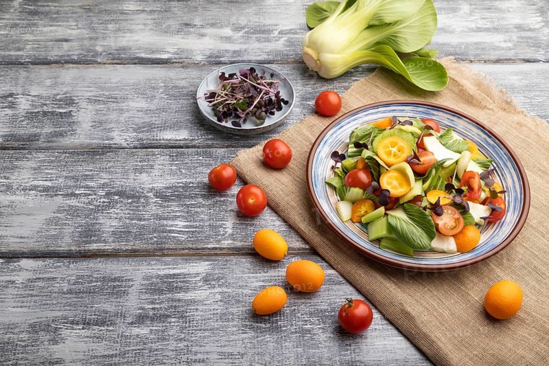 Vegetarian salad of pac choi cabbage, kiwi, tomatoes, kumquat, microgreen sprouts on gray wooden background and linen textile. Side view, copy space