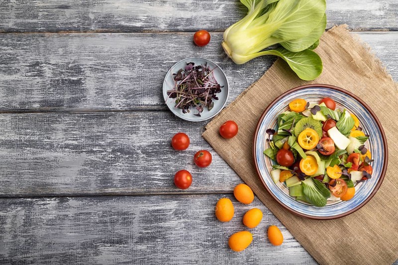 Vegetarian salad of pac choi cabbage, kiwi, tomatoes, kumquat, microgreen sprouts on gray wooden background and linen textile. Top view, flat lay, copy space