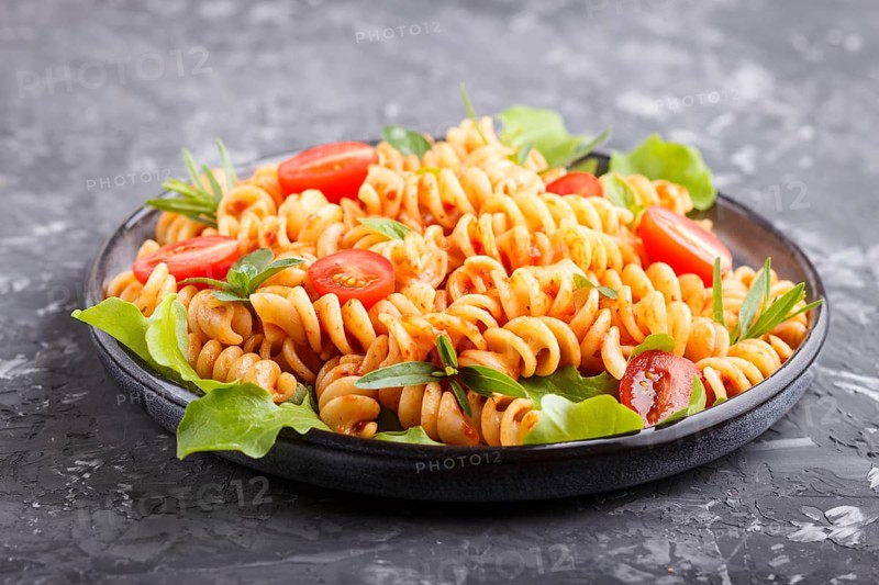 Fusilli pasta with tomato sauce, cherry tomatoes, lettuce and herbs on a black concrete background. side view, close up