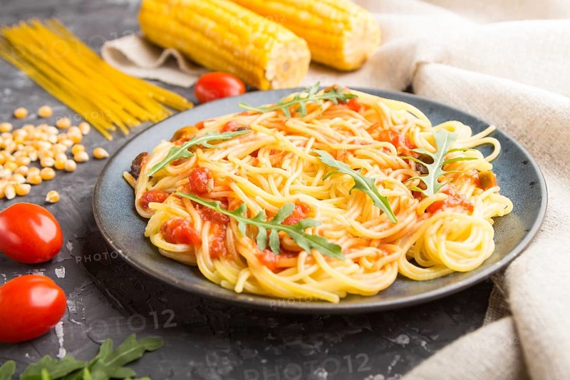 Corn noodles with tomato sauce and arugula on a black concrete background and linen textile. Side view, close up, selective focus