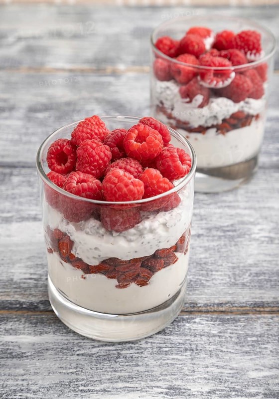 Yogurt with raspberry, goji berries and chia seeds in glass on gray wooden background. Side view, close up, selective focus