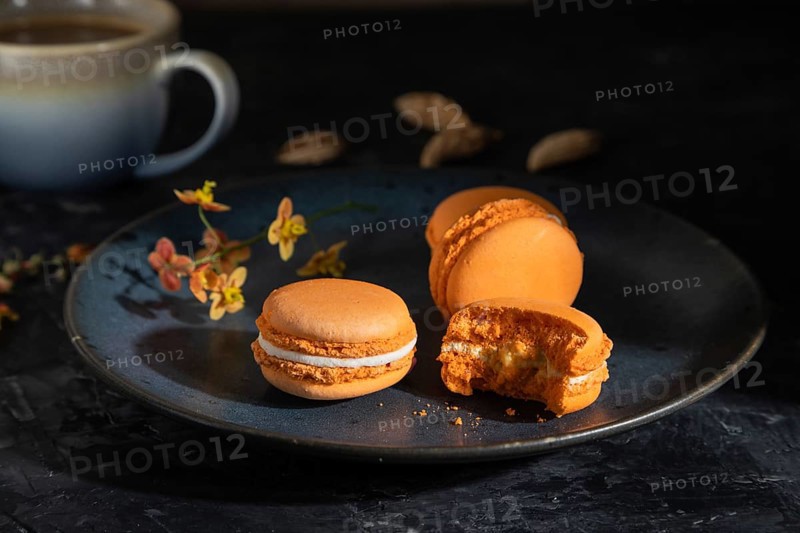 Orange macarons or macaroons cakes with cup of coffee on a black concrete background. Hard light, contrast, low key. Side view, close up