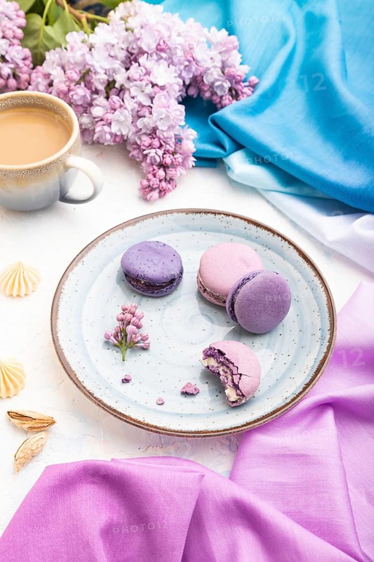 Purple macarons or macaroons cakes with cup of coffee on a white concrete background and magenta-blue textile. Side view, close up