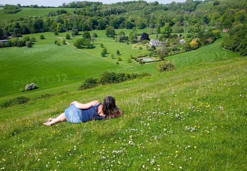 Woman lying on chalk grassland looking into deep green valley containing Rainscombe House, Oare Hill, Wiltshire, England, United Kingdom, Europe