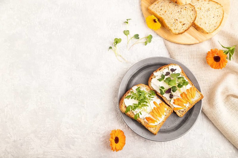 White bread sandwiches with cream cheese, calendula petals and microgreen radish and tagetes on gray concrete background and linen textile. top view, flat lay, copy space