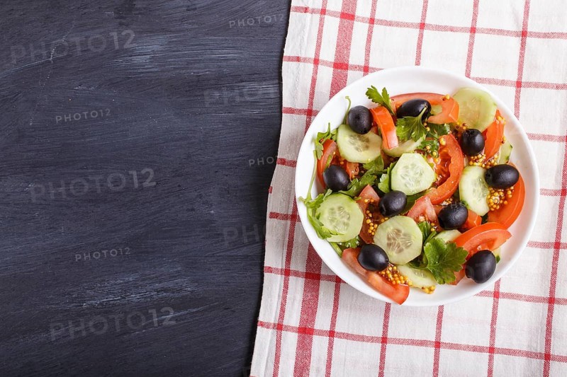 Vegetarian salad of tomatoes, cucumbers, parsley, olives and mustard on linen tablecloth and black wooden background, top view, flat lay