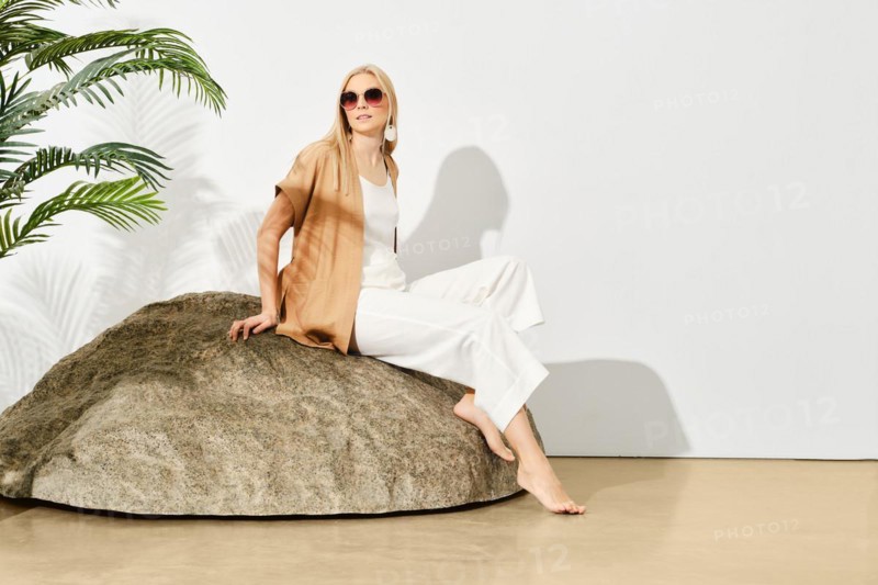 Stylish woman in a white outfit sits gracefully on a large rock inside a well-lit room filled with lush green plants, creating a serene atmosphere