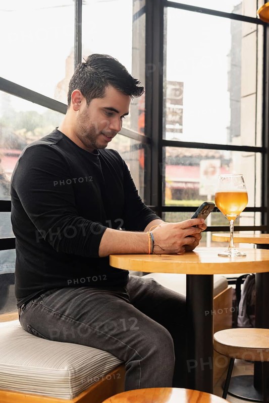 Young Latino man sitting at a bar table, using his cell phone while enjoying a glass of craft beer. The relaxed atmosphere and modern bar setting highlight a casual and enjoyable moment