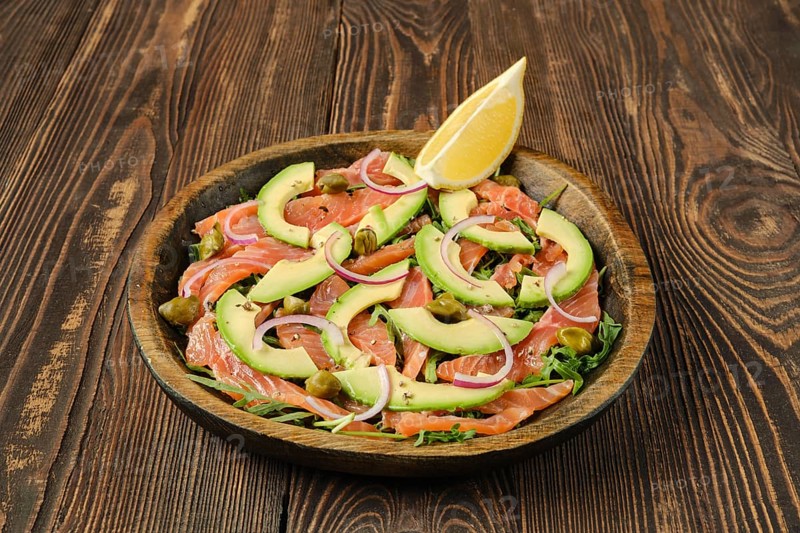 A beautifully arranged plate features slices of salmon topped with avocado, red onion, and capers, garnished with a lemon wedge, set on a rustic wooden table that enhances the dish's appeal